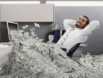 Businessman at a cubicle desk sitting in a pile of cash.